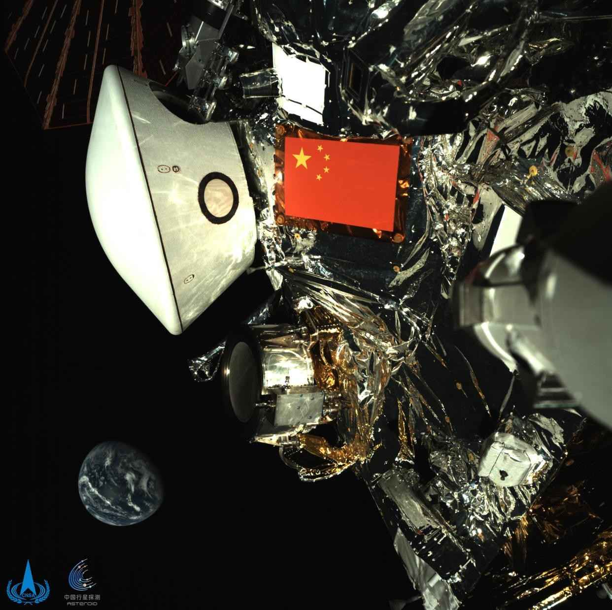 Esta imagen, publicada por la Administración Nacional del Espacio de China el 1 de octubre de 2025, muestra una vista de la sonda Tianwen-2 junto a la Tierra, captada por la sonda durante su viaje al espacio profundo. La imagen recién publicada, adquirida por una cámara de monitoreo instalada en el brazo robótico de la sonda, muestra la bandera roja de cinco estrellas de China y la cápsula de retorno blanca sobre el fondo de una Tierra azul distante. (Foto proporcionada por la Administración Nacional del Espacio de China)